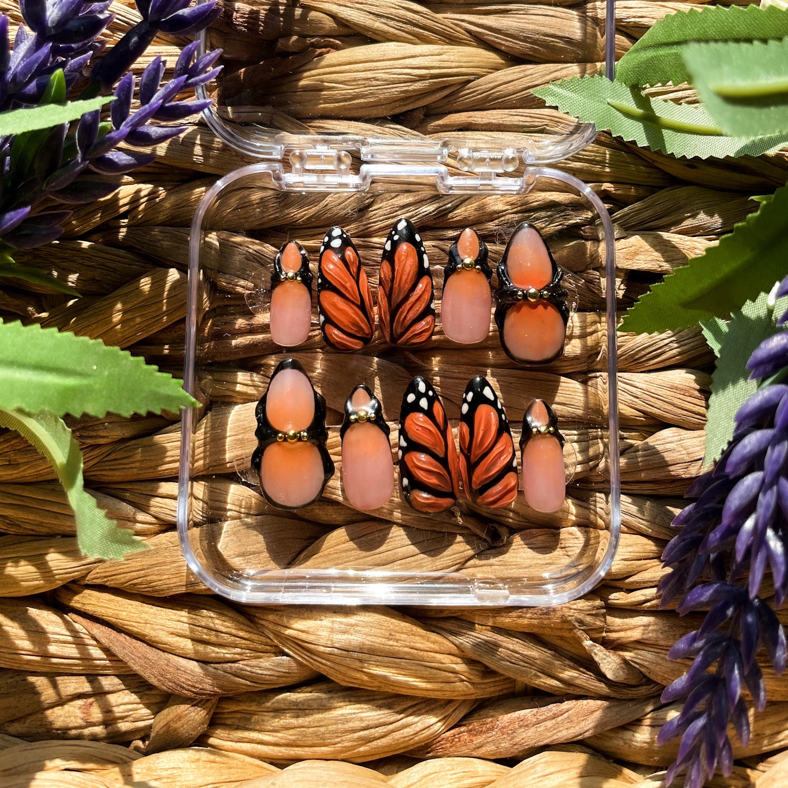 A set of press-on nails displayed in a clear case, featuring intricate monarch butterfly wing designs in orange, black, and white, with soft peach-toned accent nails, placed on a woven surface with purple and green foliage around."