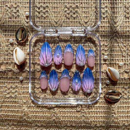 Set of custom press-on nails in a seashell-inspired design, featuring a blue-to-pink gradient with pearl and gem accents, displayed in a clear case on a woven background with seashells.