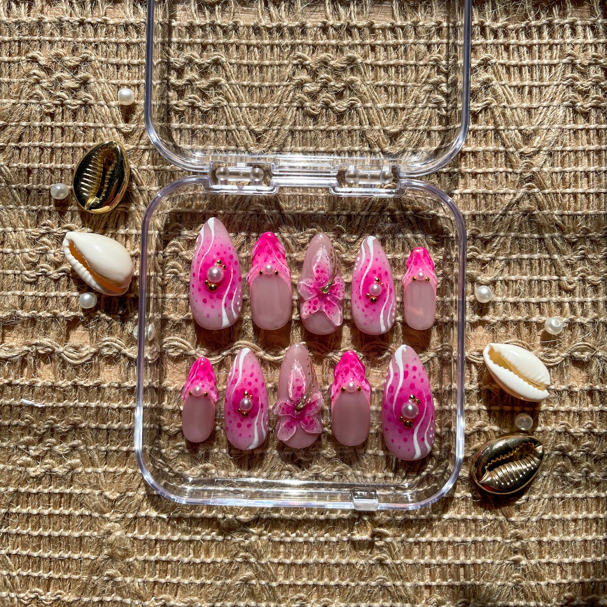 Set of press-on nails in bright pink and nude tones with pearl accents, floral 3D designs, and white wave patterns, displayed in a clear case on a woven surface with seashells and pearls.