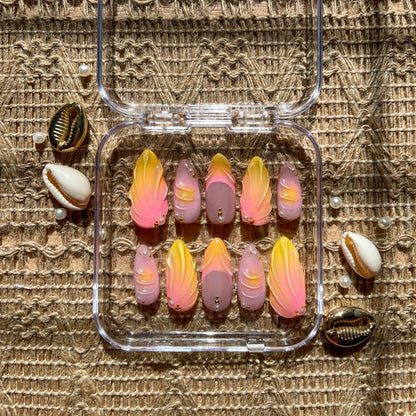 Set of press-on nails in a clear case with pink and yellow gradient designs, gold bead accents, and seashell decorations on a woven background.