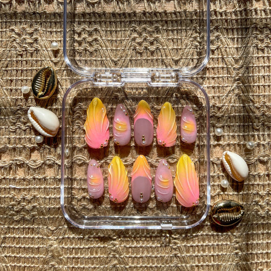 Set of press-on nails in a clear case with pink and yellow gradient designs, gold bead accents, and seashell decorations on a woven background.